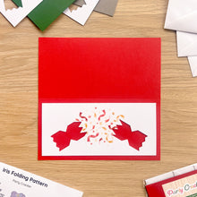 Load image into Gallery viewer, A red and white cracker aperture card and red card blank on a wooden surface, with a laser-cut red cracker bursting confetti on the white inner panel. Partial product packaging and pattern sheets are visible around it on a wood tabletop.