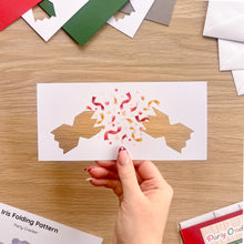 Load image into Gallery viewer, A hand holds a aperture card with a party cracker design, showing a cut-out of two half crackers with colourful confetti. The background is a wooden table with other card materials visible.