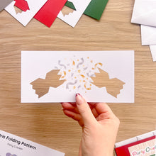 Load image into Gallery viewer, A hand holds a white rectangular card with a festive party cracker cut-out design featuring printed confetti. The background is a wooden table with colourful card blanks and white envelopes.