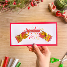 Load image into Gallery viewer, A hand holds a handmade iris fold Christmas card with a red border, featuring two red and gold cracker halves and a banner that reads 'Happy Christmas'. The card is photographed on a light wood desk with holiday decorations in the background.