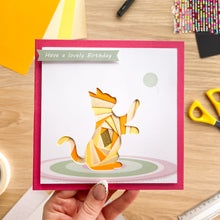 Load image into Gallery viewer, A handmade square greeting card held by a hand, featuring an iris fold cat made of orange, yellow, and white paper layers. The card has a bright pink border, a white center, a green banner that reads 'Have a lovely Birthday', and is set against a wooden desk background with craft supplies.