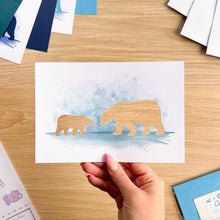 Load image into Gallery viewer, A hand holds a watercolor art print featuring the apertures of a mother polar bear and her cub against a soft blue watercolor background, set on a light wood desk with other craft materials around it.