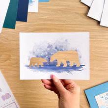 Load image into Gallery viewer, A hand holds a white art print featuring watercolor polar bear silhouettes in shades of blue and purple, with cutout bear shapes. The print is photographed from above on a light wood desk with other craft or stationery items partially visible around the edges.