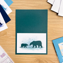 Load image into Gallery viewer, An aperture cut-out featuring a mother bear and cub on a white background, mounted on a dark teal card, displayed on a light wood tabletop with other materials around it.