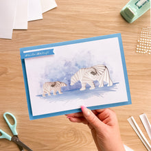 Load image into Gallery viewer, A hand holds a handmade greeting card featuring two white paper polar bears layered over a blue watercolor background. The card has a blue border and a blue banner with the text 'all is calm, all is bright'.
