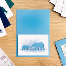 Load image into Gallery viewer, A blue greeting card with a polar bear cut-out, displayed on a light wood tabletop with other cards and stationery around it.