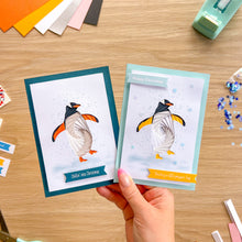 Load image into Gallery viewer, A hand holds two handmade Christmas greeting cards with penguin illustrations on a wooden craft table, surrounded by cardstock sheets, a punch tool, and glitter.