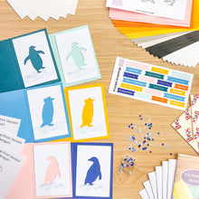 Load image into Gallery viewer, A flat-lay display of a card making kit on a light wood background, featuring colorful folded greeting cards with penguin illustrations, paper stacks, greeting sheets, iris folding guide, snowflake sequins, and other crafting supplies.