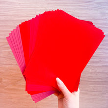 Load image into Gallery viewer, Hand holding a stack of red paper sheets on a wooden surface