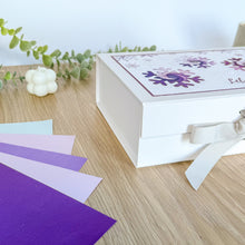 Load image into Gallery viewer, White gift box with decorative elements on a wooden surface with purple and white ribbons.