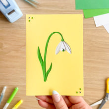 Load image into Gallery viewer, Handmade card with an iris fold snowdrop design held by a hand on a wooden surface with stationery items.
