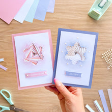 Load image into Gallery viewer, A hand holds two handmade greeting cards over a wooden craft table. One card is light pink with a layered fan design and the text 'To Someone Special'; the other is light blue with a layered fan design and 'Let it Snow'.