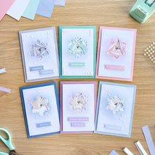 Load image into Gallery viewer, Six handmade iris fold greeting cards with snowflake designs are arranged in two rows on a wooden table, surrounded by crafting tools like scissors, a punch, and paper scraps.