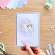 Load image into Gallery viewer, A hand holds a handmade layered Christmas greeting card with a geometric paper snowflake design and 'Happy Christmas' banner, set against a wooden craft table with stationery tools.