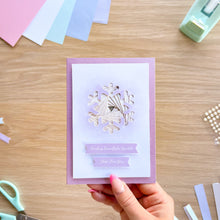 Load image into Gallery viewer, A handmade greeting card with a lavender border, a white center, and a layered die-cut snowflake design in silver and lavender. The card is held by a hand with pink manicured nails over a wooden desk background with crafting supplies.