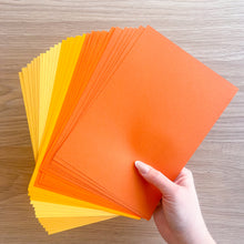 Load image into Gallery viewer, Hand holding a stack of orange and yellow paper sheets on a wooden surface