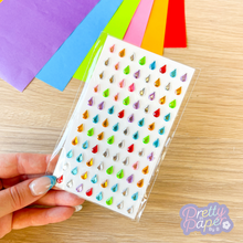Load image into Gallery viewer, Hand holding a sheet of colorful water droplet stickers with a wooden surface and colored paper in the background.