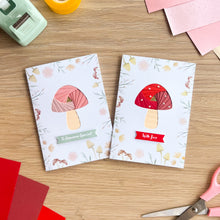Load image into Gallery viewer, Two greeting cards with mushroom designs on a wooden surface with craft supplies.