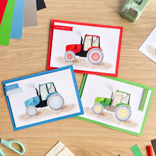 Load image into Gallery viewer, Three handmade iris fold greeting cards featuring watercolor illustrations of tractors, arranged on a light wood tabletop with craft supplies nearby. The cards have red, blue, and green borders and display tractors in red, blue, and green respectively.