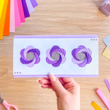 Load image into Gallery viewer, Hand holding a card with purple flower designs on a wooden table with craft supplies.