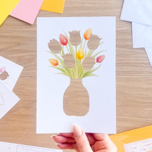 Load image into Gallery viewer, Hand holding a card with a cut-out vase of tulips on a wooden surface.