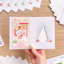 Load image into Gallery viewer, A hand holds a packaged Baby Gonk card aperture pack on a light wood tabletop, with printed cards and white sheets arranged around it.
