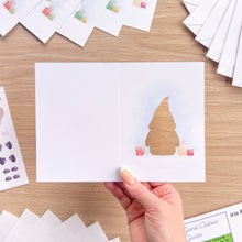 Load image into Gallery viewer, A hand holds an open white greeting card on a light wood table. The right panel shows a cut-out of a small gonk, surrounded by pastel gift boxes and a faint blue watercolor wash.
