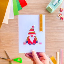 Load image into Gallery viewer, A hand holds a white greeting card featuring a cute iris fold Christmas gonk with a striped red and white hat, a red nose, and a red coat. The card is photographed from above on a light wood desk surrounded by craft supplies like scissors, colored paper, and a tape dispenser.
