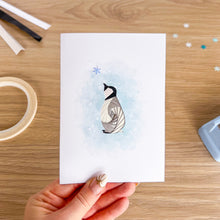 Load image into Gallery viewer, A hand holds a white greeting card featuring an iris fold penguin with a light blue watercolour background. The card is photographed from above on a wooden desk with craft supplies like tape, scissors, and glitter scattered around.
