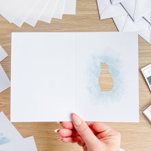 Load image into Gallery viewer, Hand holding a card with a watercolor design of a penguin on a wooden table.
