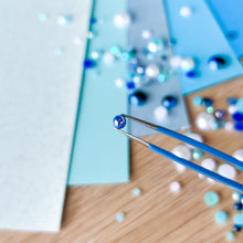 Load image into Gallery viewer, Close-up of tweezers with a blue gemstone on a wooden surface with scattered gems.
