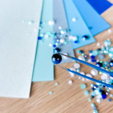 Load image into Gallery viewer, Blue tweezers picking up small blue and white beads on a wooden surface with blue paper.
