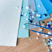 Load image into Gallery viewer, Close-up of tweezers holding a small gemstone with a blurred background of more gems and materials.
