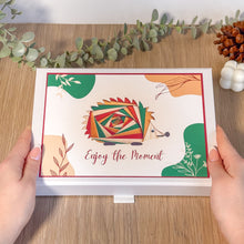 Load image into Gallery viewer, Colourful hedgehog illustration on a gift box held by hands with decorative elements on a wooden surface.
