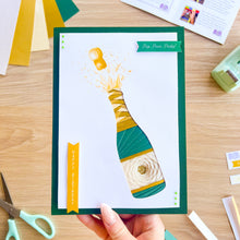 Load image into Gallery viewer, Handmade card with a champagne bottle design held by a hand on a wooden table with crafting supplies.
