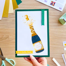 Load image into Gallery viewer, Hand holding a greeting card with a champagne bottle design on a wooden surface.
