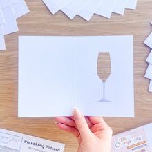 Load image into Gallery viewer, Hand holding a scored card blank with a champagne glass cut-out with printed silver stem on a wooden surface
