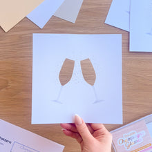 Load image into Gallery viewer, Hand holding a paper cut-out of two champagne glasses on a wooden surface with coloured card in the background.
