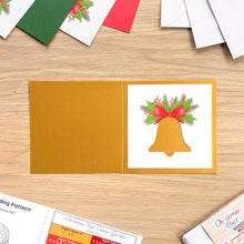 Load image into Gallery viewer, A Christmas bell card is on a light wood tabletop. The left panel is a warm mustard-gold color, and the right panel shows a white background with a gold bell illustration topped with green holly and a red bow.
