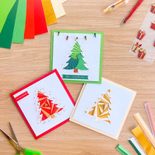 Load image into Gallery viewer, Three handmade geometric Christmas greeting cards arranged on a light wood desk, surrounded by craft scissors, colored paper strips, and small wrapped gift decorations. The cards feature stylized tree designs in red, green, and gold with festive text.
