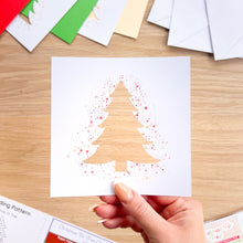 Load image into Gallery viewer, A hand holds a square white card featuring a Christmas tree silhouette made from red and gold glitter or paint splatter, with other craft cards and envelopes scattered on a light wood tabletop.
