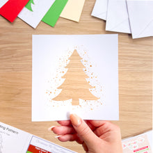 Load image into Gallery viewer, A hand holds a white square card featuring a laser-cut Christmas tree design with gold glitter splatter details, set against a wooden tabletop with craft supplies in the background.
