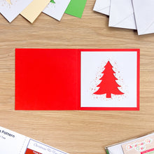 Load image into Gallery viewer, An open red greeting card laid on a light wood tabletop, with a white insert panel featuring a red Christmas tree silhouette and red paint splatter details. Partially visible cards, envelopes, and printed pattern sheets surround it on the tabletop.
