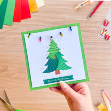 Load image into Gallery viewer, A hand holds a square handmade Christmas card with a green border and a green iris fold tree design, set against a wooden craft table with colorful paper strips and small gift decorations.
