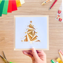 Load image into Gallery viewer, A hand holds a square greeting card featuring an iris fold Christmas fir tree design on a white background. The card is photographed from above on a light wood desk with craft supplies like colored paper, scissors, and markers visible around it.
