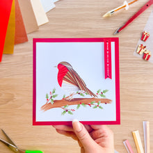 Load image into Gallery viewer, A handmade square greeting card with a red border and an illustration of a bird on a branch, held by a hand over a wooden craft desk with scissors, colored paper, and other crafting tools.

