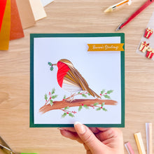 Load image into Gallery viewer, A hand holds a square greeting card featuring a red-breasted bird perched on a branch with holly leaves and berries, set against a light wood desk background with craft supplies around it.
