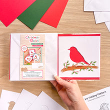 Load image into Gallery viewer, A hand holds a Christmas Robin Card Aperture Pack on a light wood table. The set includes embellished card cut-outs of a robin bird on a branch and an iris folding pattern.
