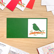 Load image into Gallery viewer, Green and white Christmas card materials featuring a bird perched on a branch, displayed on a light wood tabletop with other cards and envelopes around it.
