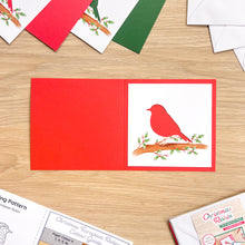 Load image into Gallery viewer, A red greeting card blank laid flat on a light wood surface, with a red bird cut-out perched on a branch printed on the white panel. Other colorful cardstock sheets and printed guides are partially visible around it.
